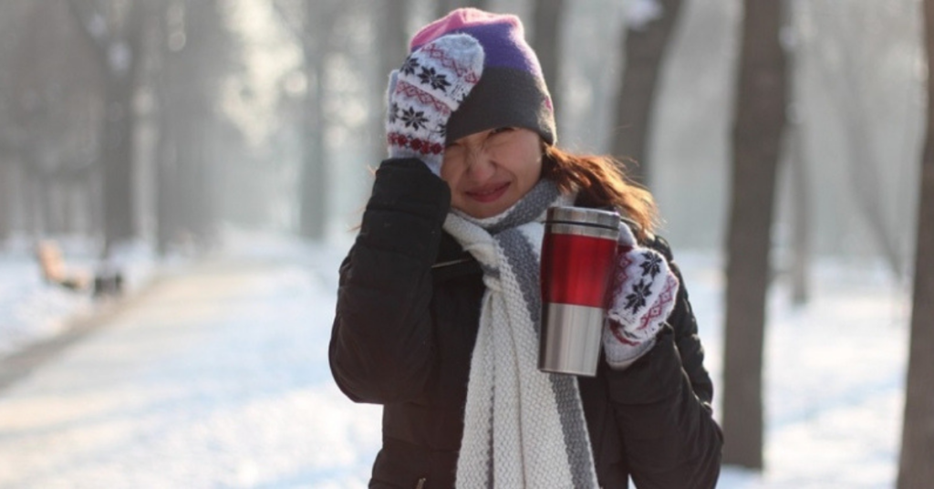 A women bundled up during a winter day with her hand on her head and holding a coffee.