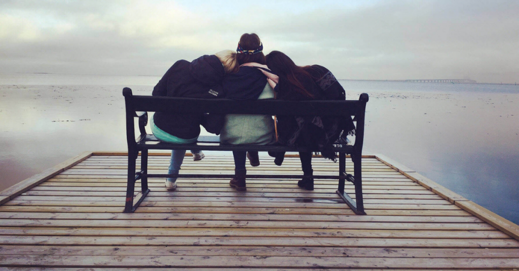 Three women sitting on a bench, leaning on each other while looking out at a lake.