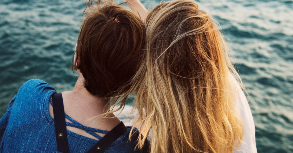 Two women sitting down and leaning on each other while they look out over the water.