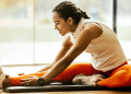 Woman in athletic wear sits on a mat, reaching for her toes in a seated forward bend during a workout indoors.