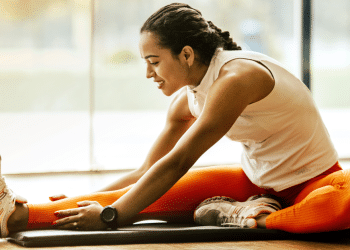 Woman in athletic wear sits on a mat, reaching for her toes in a seated forward bend during a workout indoors.