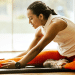 Woman in athletic wear sits on a mat, reaching for her toes in a seated forward bend during a workout indoors.