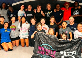 Group of young wrestlers posing in a gym, standing and kneeling together while holding a black banner with logos.