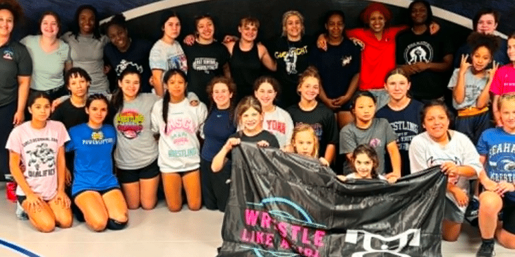 Group of young wrestlers posing in a gym, standing and kneeling together while holding a black banner with logos.