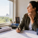 Woman architect at a sunlit desk by a window, drawing on architectural blueprints with a pen; mug reads 'MILITARY SPOUSE'.